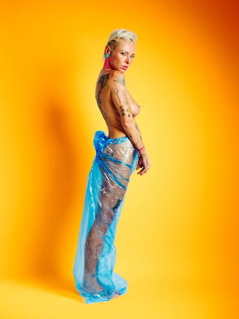 Explicit art nude portrait of Lu Epic in blue plastic pants with vivid yellow backdrop